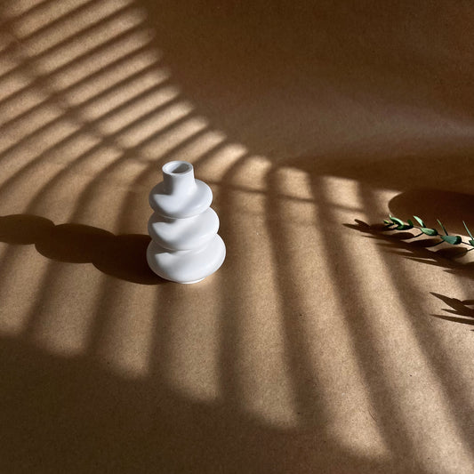 A white, handmade ceramic decorative vase with a modern geometric design, placed on a surface casting shadow patterns, likely created by the afternoon sun.